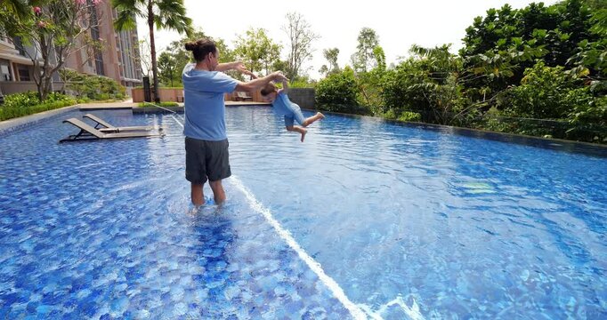Father stands in shallow part of pool, holding his toddler son's hands and spinning him around. Little one kicks his legs amusingly, suddenly father releases him, toddler joyfully flies and plunge in.