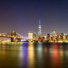 Obraz premium Illuminated cityscape at night, with a prominent bridge and numerous skyscrapers reflected in the calm water. The sky transitions to a dark hue