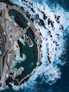 Aerial view of the natural volcanic swimming pools with crashing blue waves and white foam against dark rocks in Porto Moniz, Madeira, Portugal.