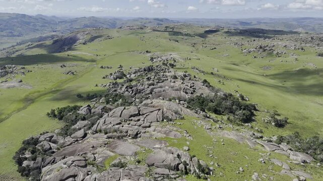 Drone flies downhill from summit toward grassy fields with boulders on a sunny day at Sibebe Rock near Mbabane, Eswatini
