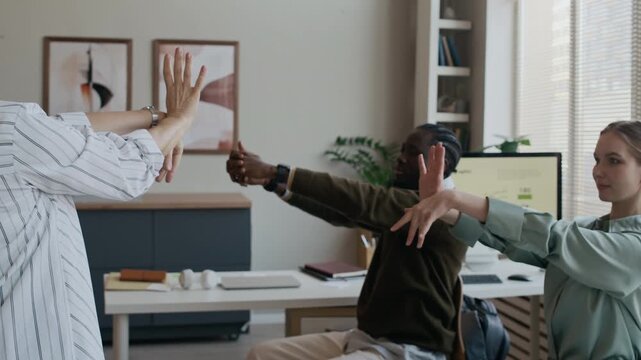 Group of diverse young adult and senior colleagues exercising during break in office, stretching their wrists and fingers, preventing carpal tunnel syndrome