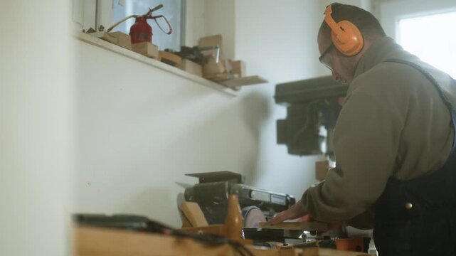 A skilled craftsman diligently sands a piece of wood in his bright workshop. The man uses a power sander while wearing protective earmuffs and safety glasses.