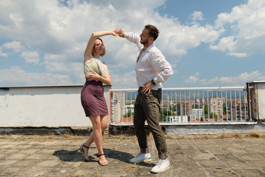 Happy young couple dancing on a rooftop terrace with panoramic city view on a sunny day. Modern romance, dating lifestyle, urban living and carefree relationship concept