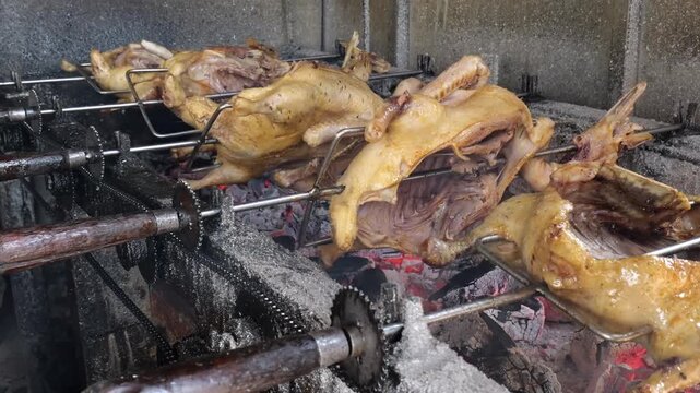 Row of whole ducks roasting on metal rotisserie skewers over glowing red charcoal embers .The golden brown poultry is being cooked traditionally with a mechanical gear system visible