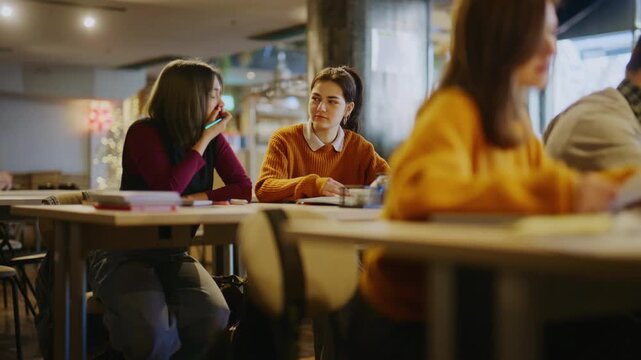 Two female students are engaged in a discussion while studying at a table in a university setting. They are collaborating on academic work.