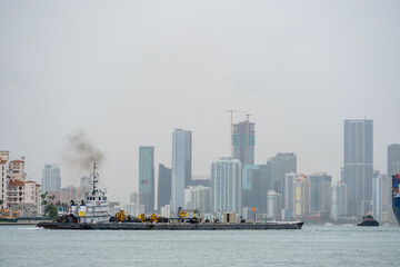 Obraz premium Industrial Tugboat Pushing Barge Near Port of Miami. Tugboat with exhaust smoke pushing a heavy cargo barge through the water.