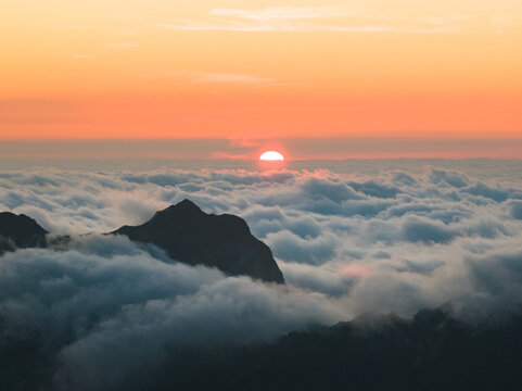 Aerial view of Pico do Arieiro mountain peak rising above a thick sea of clouds during a vibrant orange sunset in Curral das Freiras, Madeira, Portugal.