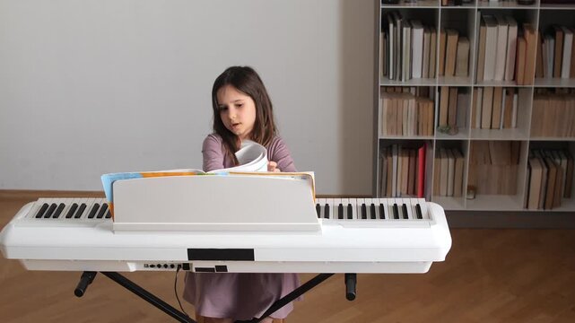 Young girl concentrating on sheet music while practicing an electronic white piano at home, focused on learning piano, developing musical skills and enjoying a quiet music lesson