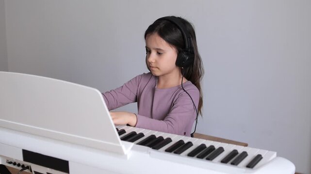 Young girl wearing headphones concentrates on playing a white digital piano during a focused music lesson or solo practice session at home, developing skills and creativity