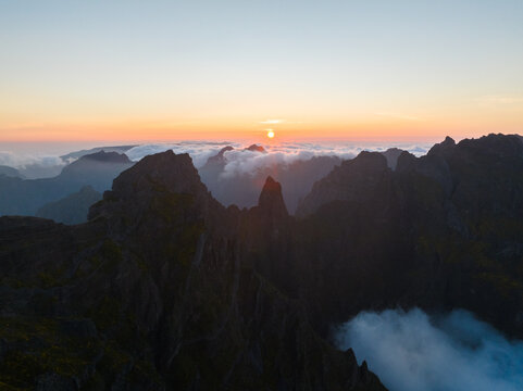 Aerial view of Pico do Arieiro with jagged mountain peaks rising above a sea of clouds during a golden sunset in Curral das Freiras, Madeira, Portugal.