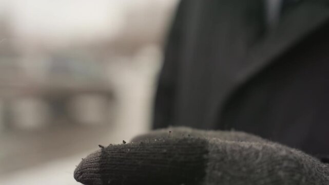 Gloved hand holding cigarette on cold street, worn wool glove with torn fingertips, ember glowing between fingers, blurred urban background, dark overcoat and tie visible, muted color palette, bleak