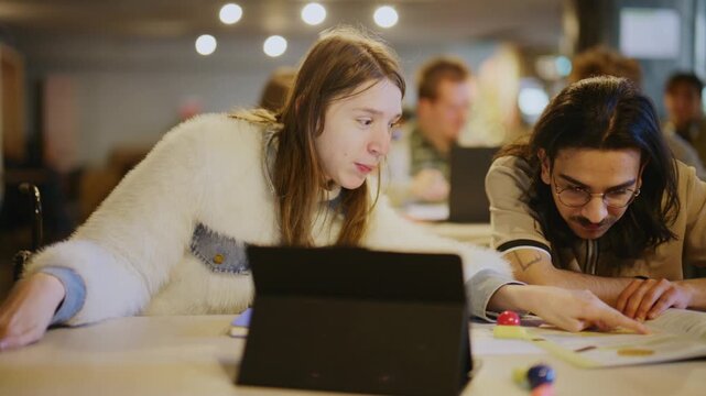 Students engage in collaborative learning, focusing on a textbook and discussing their work together. Young people are seen studying and utilizing technology in a modern educational environment.