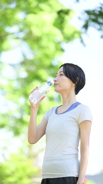 新緑の屋外で水分補給をする女性の横顔ポートレート　ウォーキングやランニングの運動やスポーツの間にペットボトルの水を飲むクローズアップ　春夏の半袖の縦動画