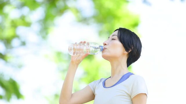 新緑の屋外で水分補給をする女性の横顔ポートレート　ウォーキングやランニングの運動やスポーツの間にペットボトルの水を飲むクローズアップ　春夏の半袖　スローモーションの動画