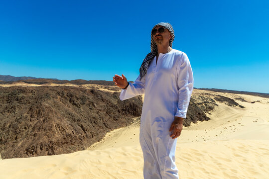 Arab man in national dress stands in the Sinai or Sahara desert and looks at the blue sky. Man in traditional thobe tobe thawb dishdasha kandura and sunglasses standing proudly in desert.