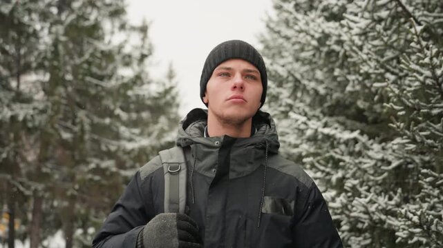 Young white man in snowy forest wearing black parka and knit beanie, backpack strap over shoulder, outdoor hiker stance, looking upward with calm determination, frosted pine trees and falling snow,