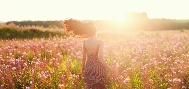 Young woman wearing open dress running with flying hair through blooming flower field at back light sunset. Spring and summer fashion. Rear view.