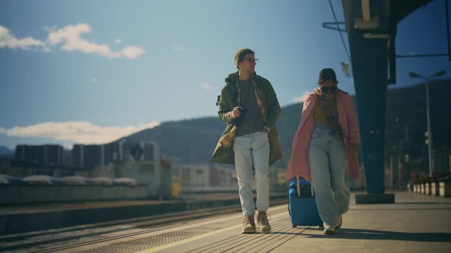 A young couple of tourists walks along a train platform, pulling their luggage behind them. They appear ready to embark on a journey in an urban setting.