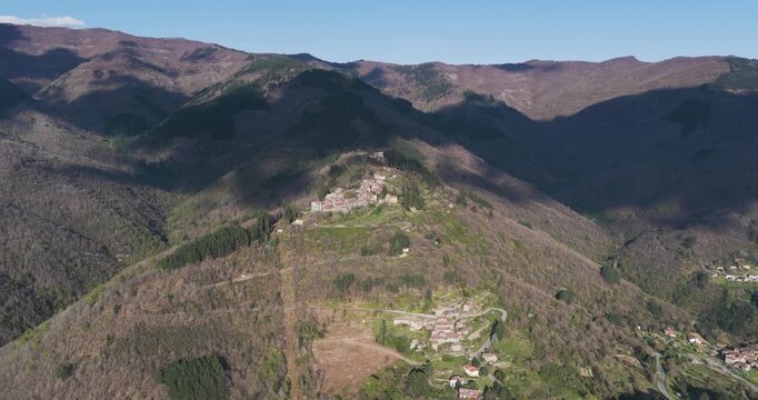 Aerial view of the small village of Trappola nestled on a steep forested hillside with mountain peaks and cloud shadows in Trappola, Tuscany, Italy.