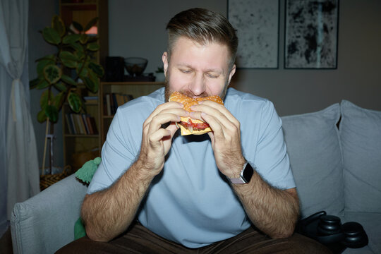Caucasian young adult man eating burger while sitting at home, showing fast food craving and unhealthy diet choice. Useful for nutrition campaigns, obesity risk, lifestyle marketing