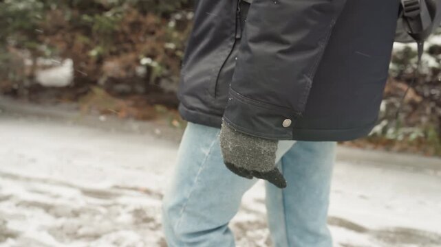 Lower body of white man walking on snowy sidewalk, blue jeans and wool mittens visible, black parka and backpack edge, footprints and slush on pavement, casual streetwear winter vibe with muted tones