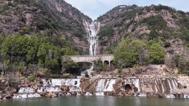 Aerial drone view of Tiantai Mountain waterfall tumbles over rugged cliffs into peaceful lake, spanned by an elegant stone arch bridge and framed by lush green foliage under clear skies