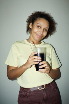 Portrait of young adult Black woman holding glass of soda and smiling at camera against plain wall. Useful for beverage advertising, refreshment campaigns, vertical