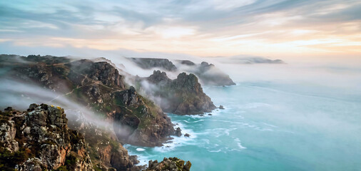 Fototapeta premium Coastal cliffs shrouded in atmospheric mist at sunrise with beautiful turquoise ocean waters and dramatic cloudy sky landscape, showcasing natural beauty.