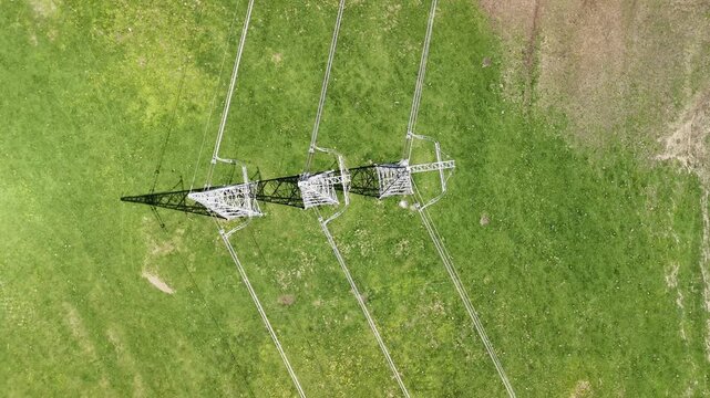 High voltage electricity power line tower with cables and insulators transmitting energy through a green meadow, an aerial top down view of the transmission pylon