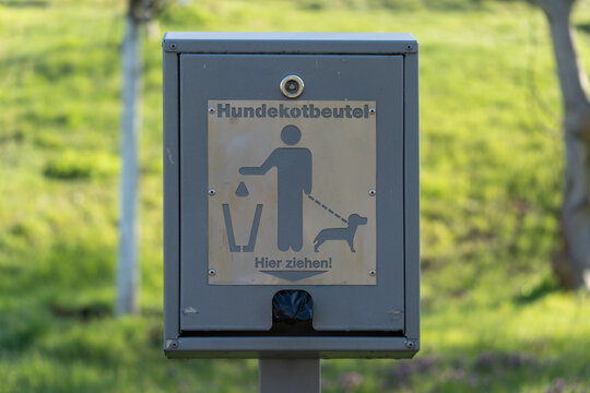 Gray metal box for dog waste disposal. Sign shows person with leash and dog. Text reads Hundekatobutel and Hier ziehen. Green grass and trees blur softly behind. Ideal for parks or public areas