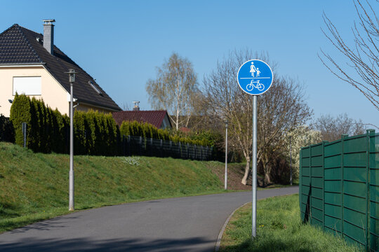 Blue bicycle sign stands beside quiet village road. Green fence lines the roadside with trimmed hedges. Sloping path leads toward residential homes under clear sky