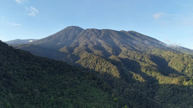 A view of lush green mountains with dense tropical forests and soft clouds drifting along the slopes under a clear blue sky. Mount Gede Pangrango, Puncak Bogor.