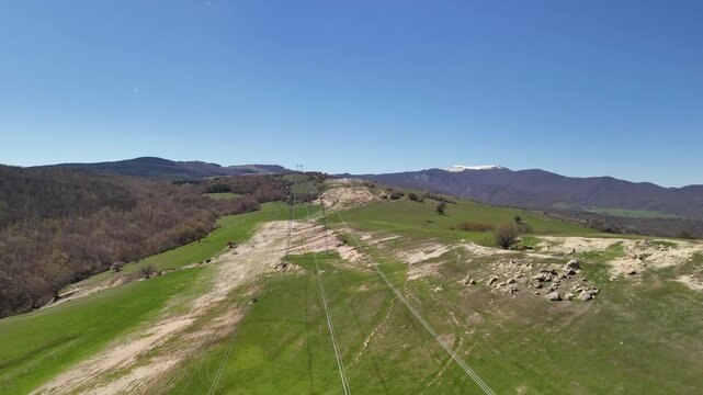 Aerial backwards flight revealing power lines cutting through a vibrant green mountain valley, with a snow capped peak and clear blue sky in the background