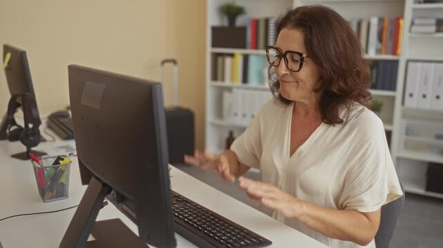 Woman at computer raises arms at desk in a building, smiling and cheering with glasses, keyboard and bookshelves visible; joy success celebration.