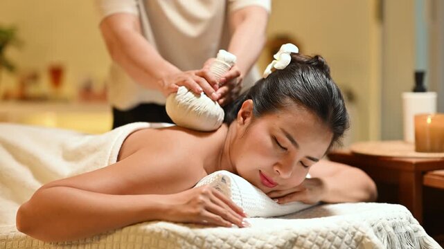 Woman receiving herbal compress massage treatment at spa, therapist applying hot compress on back in warm relaxing atmosphere.
