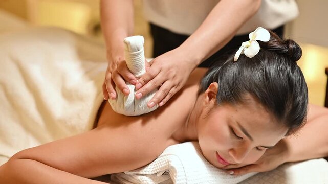 Woman receiving herbal compress massage treatment at spa, therapist applying hot compress on back in warm relaxing atmosphere.