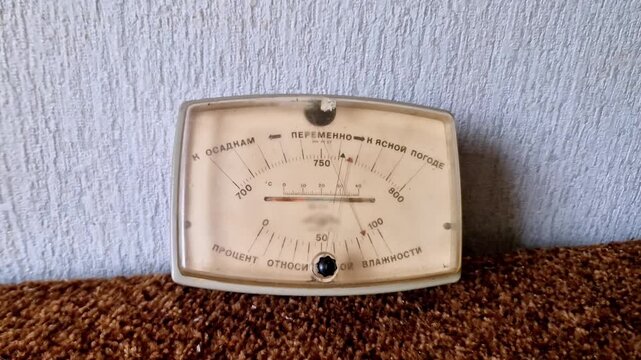 Close-up of a vintage analog barometer with Cyrillic text resting on a brown textured carpet. The needle indicates changeable weather conditions against a light blue wall background.