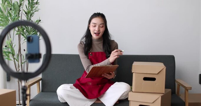 Asian woman livestreaming fashion from a cozy home studio, promoting her small ecommerce boutique on social media using smartphone and ring light to showcase clothes and connect with customers