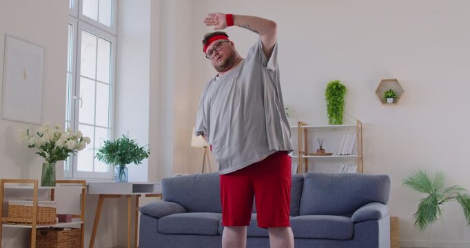 Funny plus size man doing side bends at home. Obese person in sportswear stretching and warmup training, morning weight loss workout and physical exercises of chubby guy with obesity in living room