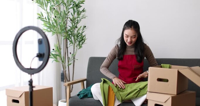 Asian woman livestreaming fashion from a cozy home studio, promoting her small ecommerce boutique on social media using smartphone and ring light to showcase clothes and connect with customers