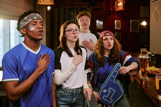 Multiethnic young adult sports fans singing anthem in bar, supporting team during match viewing. Ideal for fan culture, group loyalty, sports marketing, medium shot