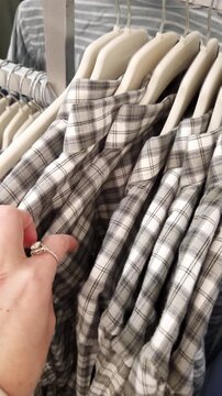 Female hand with rings slides across a row of grey and white plaid flannel shirts. Close-up shot captures the texture of the fabric and the experience of shopping in a store.