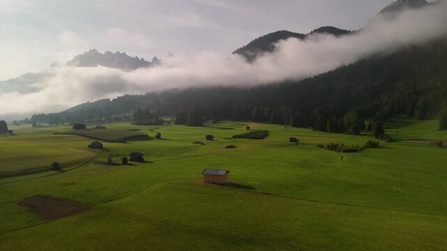 A typical wooden cabin stands in a meadow in the Bolzano region of South Tyrol. In the background rise the majestic mountains of the Sexten Dolomites, shrouded in clouds. drone perspective.