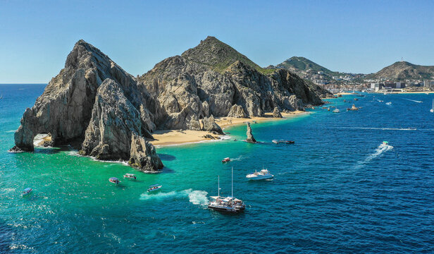 Aerial view of the iconic El Arco rock formation meeting the turquoise sea, boats dotting the water under a clear sky, Cabo San Lucas, Mexico.
