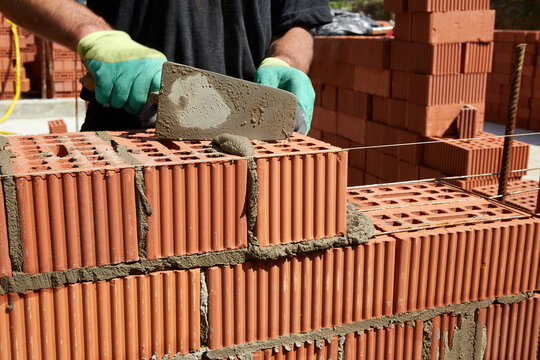A skilled bricklayer is carefully applying mortar to red bricks, aligning them precisely to build a wall at a construction site