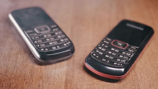 Two black vintage mobile phones with physical keypads lie side by side on a textured wooden table. Concept of obsolete technology, communication evolution, and the simplicity of retro devices