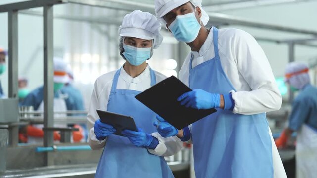 Two quality control specialists in sanitized uniforms and masks collaborating on production data and audits in a modern industrial food processing facility.