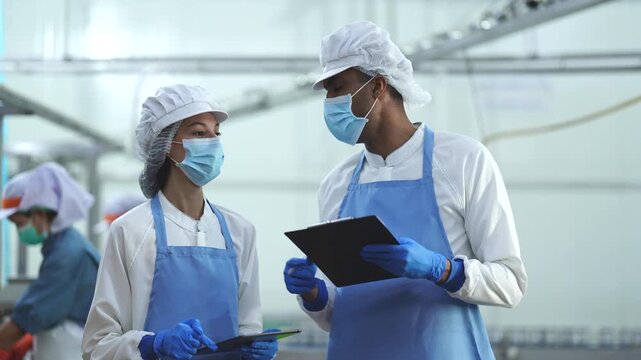 Two quality control specialists in sanitized uniforms and masks collaborating on production data and audits in a modern industrial food processing facility.
