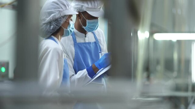 Two quality control specialists in sanitized uniforms and masks collaborating on production data and audits in a modern industrial food processing facility.