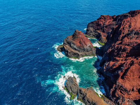 Aerial view of the rugged, red volcanic cliffs meet the turquoise ocean as waves crash against the shore, Capelo, Acores, Portugal.